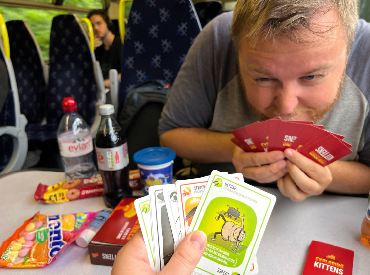 Eit bilete teke over eit bord om bord på eit ScotRail-tog på veg frå Glasgow til Fort William. I framgrunnen held ei hand ei vifte av kvite kort frå kortspelet «Exploding Kittens». Det fremste er eit grønt «Defuse»-kort med teksten «via participation in kitten yoga» og ei teikning av ein naken mann som gjer yoga saman med ei katt, og bak det skimtar ein eit oransje «Attack»-kort. På andre sida av bordet sit ein mann med skjegg og grå t-skjorte. Han lener seg fram, held dei raude korta sine opp mot nasen og skeiar lurt mot kameraet med eit lite glis. Bordet flyt over av reisesnop: ei flaske Evian, ei flaske Diet Coke, eit boks Mini Oreo med blått lok, ein pakke Jammie Dodgers med prislappen «£1.29», ein pose Fruit Mentos, ein flaske handsprit og den raude spelesken. I bakgrunnen står mørkeblå seterader med kvitt kryssmønster og gule handtak, ein ung mann med mørkt hår sit lenger bak og ser mot kameraet, og gjennom vindauget glid grøne tre forbi.